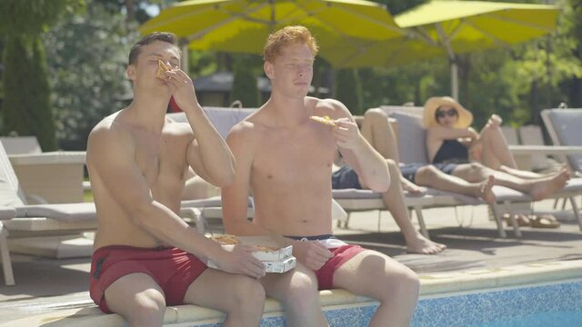 Two young Caucasian men sitting at poolside and eating tasty pizza. Portrait of happy relaxed male friends resting at summer resort on sunny day. Tourists enjoying vacations. Tourism concept.
