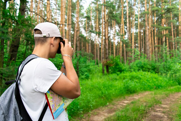 Back view of a male tourist in a white t-shirt cap with a gray backpack, walking along a path in the forest, turning his head.Tourist with a map and camera.