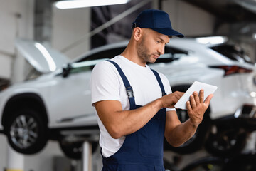 handsome mechanic in overalls and cap using digital tablet in car service
