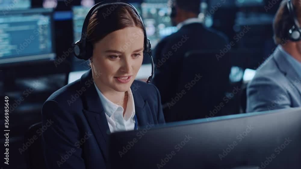 Poster Happy Beautiful Technical Customer Support Specialist is Talking on a Headset while Working on a Computer in a Dark Monitoring and Control Room Filled with Colleagues and Display Screens.