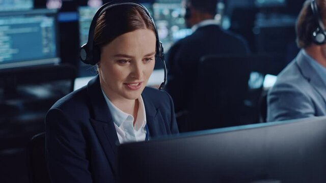 Happy Beautiful Technical Customer Support Specialist is Talking on a Headset while Working on a Computer in a Dark Monitoring and Control Room Filled with Colleagues and Display Screens.