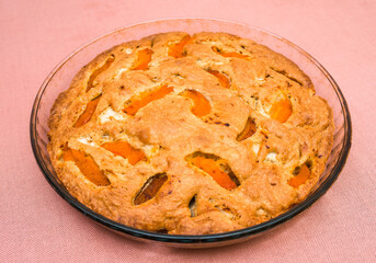 A large, round baked apple and apricot pie stands in a glass form on a table with a tablecloth.