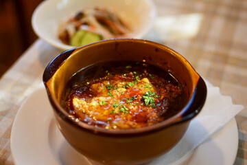 陶器の茶色い器に盛られた、オニオングラタンスープ。日の光。背景にサラダ。レストランのテーブル。太陽光。