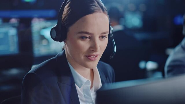 Close Up Portrait of a Happy Beautiful Technical Customer Support Specialist Talking on a Headset while Working on a Computer in a Dark Monitoring Room Filled with Colleagues and Display Screens.