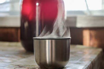 Clear steam over a thermos cup on a background of the jar with red homemade wine on a cold sunny day.  Evaporation of liquid. Copy space. 