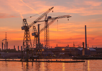 Obraz premium Tower cranes and elements of an oil refinery are silhouetted against a brilliant sunset near Antwerpn, Belgium.