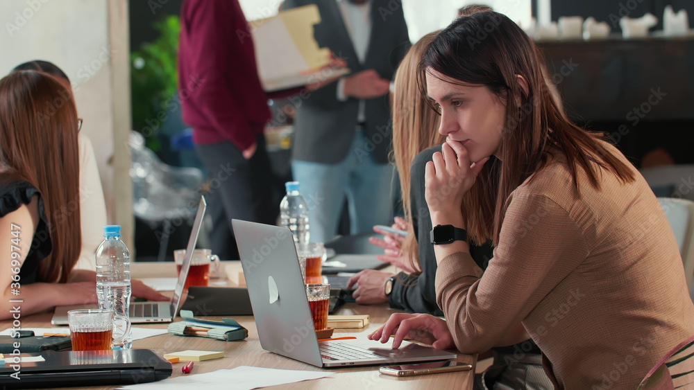 Sticker Side view beautiful focused young Caucasian woman using laptop working at busy coworking table full of business people.