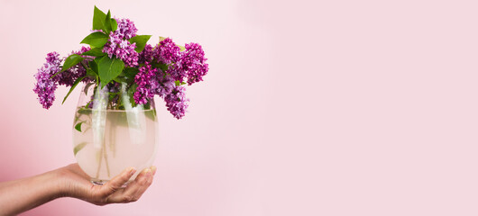 girl holds a bouquet of lilacs in a vase on a pastel pink background