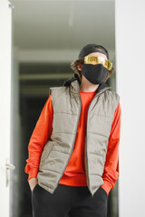 A young man of 25-30 years old in a black protective mask, yellow goggles, a cap and a red jacket posing on a gray background.