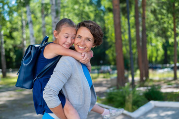 Naklejka premium A beautiful caucasian woman stands outdoors and holds daughter on her back. A cheerful smiling girl sits on her mother back while walking in the park. Friendly loving family.