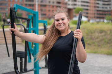 Beautiful smiling overweight young woman does fitness on an ellipsoid simulator outdoors. Fat girl is training on the sports ground for weight loss. Sport outside on a warm summer day.