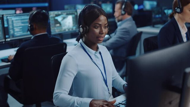Joyful Beautiful Technical Customer Support Specialist is Talking on a Headset while Working on a Computer in a Call Center Control Room Filled with Colleagues, Display Screens and Data Servers.