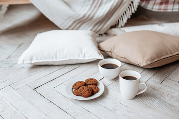 Breakfast in the morning. Two white cups of coffee and chocolate chip cookies stand on a light wooden parquet and pillows lie nearby. Homeliness, day off. Pastel colors.