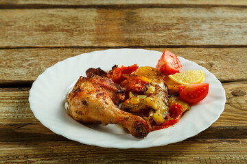 Baked chicken leg with potatoes and tomato on a white plate on a wooden table.
