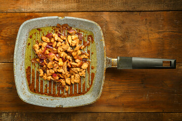 Frying pan with fried chicken fillet and onions on a wooden table.