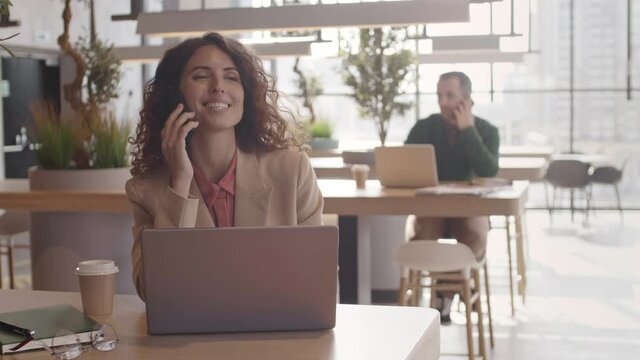 Medium shot portrait of beautiful curly-haired businesswoman sitting at table in coworking cafe, finishing phone conversation and looking at camera while man working on background