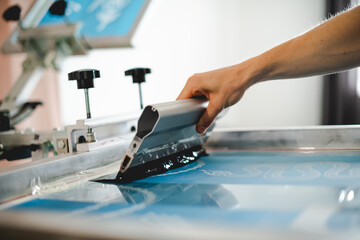Cropped view of artisan holding squeegee with plastisol ink while working with screen printing machine in designer studio  - obrazy, fototapety, plakaty