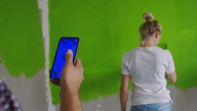 A man's hand swipes across the phone screen with a chromakey, and a girl paints the wall in the background. Close-up of a man's hand with a phone with a blue screen, against which a girl paints the