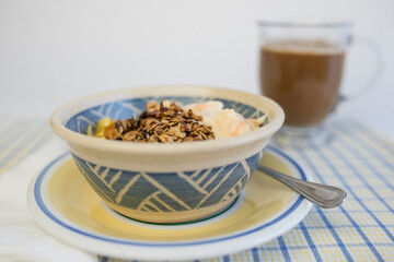 Homemade oat and nut granola in decorated ceramic bowl with peach yogurt and glass of coffee in the background.