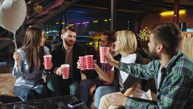 In a modern stylish loft apartment in living room young group of friends celebrating a birthday cheers with color cups and enjoying the time together