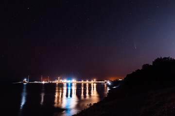 Comet C/2020 NEOWISE at night star sky and sea with reflection of city lights