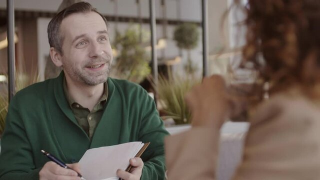 Back view of Caucasian middle-aged man sitting at cafe table, making notes in notepad while interviewing curly-haired unrecognizable woman sitting in front of him