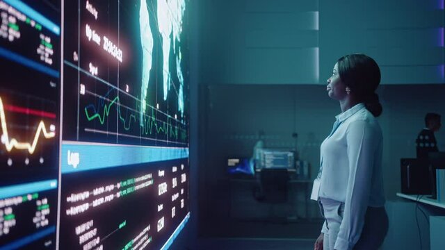 Young Female Computer Science Engineer Looking at Big Screen Display Showing Global Map with Data Points. Telecommunications Technology Company System Control and Monitoring Room with Servers.