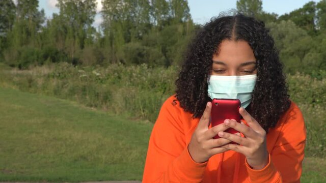 Beautiful mixed race African American girl teenager young woman wearing a face mask during COVID-19 Coronavirus pandemic using her smartphone or cell phone outdoors for social media or text messaging