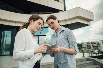Selective focus of two young female colleagues standing near glass office building and watching information in white tablet pc. Business concept. Confident ladies with modern device.