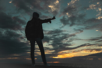 Silhouette of a man on the ocean in front of a strewn sky with colored clouds shows with his hand...