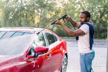 African stylish young focused man in casual clothes washing the red car with a water gun on...