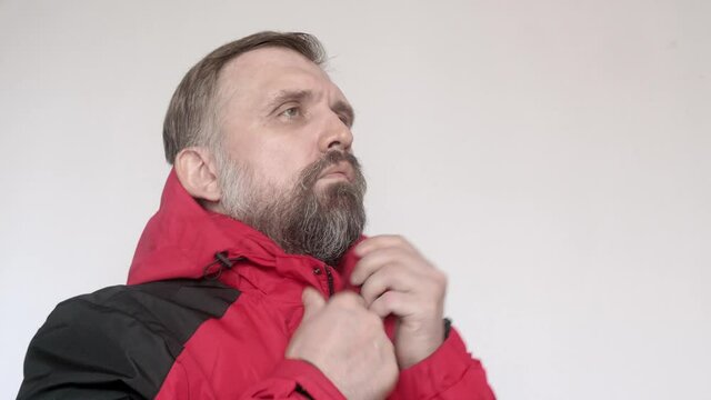 Man with white wall behind him puts on red coat and black baseball cap.