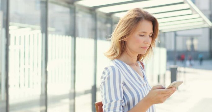 Caucasian young beautiful blonde woman using smartphone outdoor on sunny day. Pretty female texting message on cellphone at bus stop. Lady tapping and scrolling on mobile phone, waiting for transport.