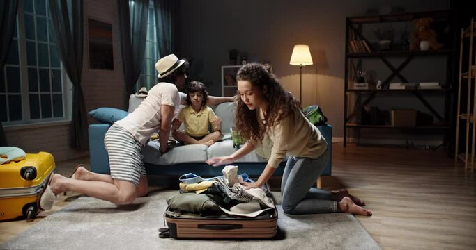 Funny asian kid with curly hair meditating on the couch while his young parents are rapidly packing clothes into suitcase for a flight. Happy family preparing for adventure 4k footage