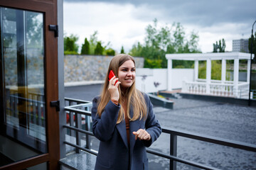 Fototapeta premium Young beautiful blonde woman in gray coat talking on the phone on a cold autumn day. Girl with phone on the terrace overlooking the river