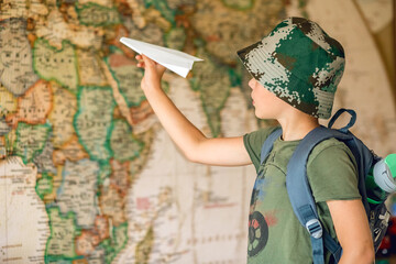 Fototapeta na wymiar A child with a backpack on his back and a paper plane in his hand dreams of becoming a traveler. The back blurred background is a map of the world.