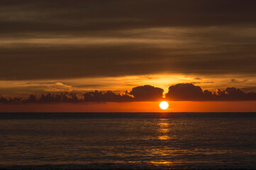 Sun rising from the sea between two clouds and the sea with soft waves bathing the shore of the beach behind the clouds