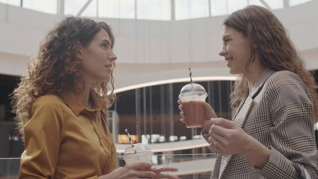Back view of two young Caucasian women wearing casual clothes standing in store with beverages in their hands and having communication