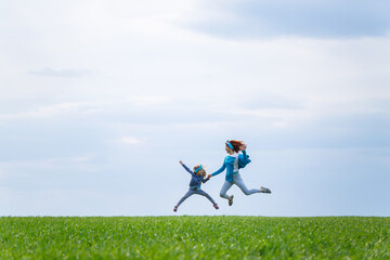 little girl child and mother woman run and jump, green grass in the field, sunny spring weather, smile and joy of the child, blue sky with clouds