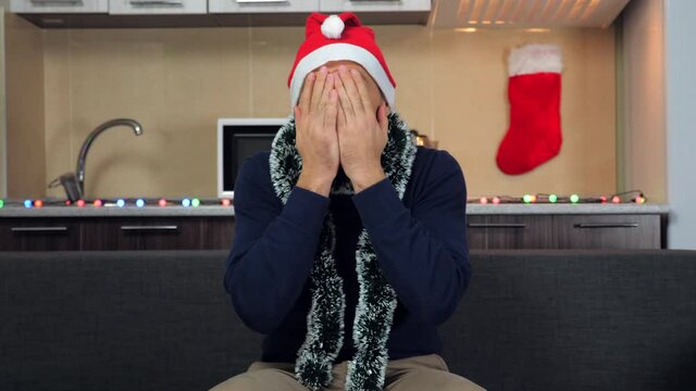 Shocked man in New Year red Santa Claus hat watches television covers face with hands. Guy in blue sweater sits couch at home on background Christmas kitchen, afraid watch TV, covers mouth, eyes palms
