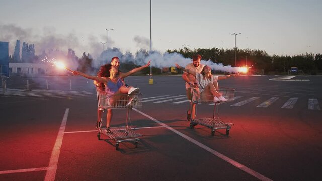 Guys riding and spinning on shopping trolleys with their girlfriends at deserted parking lot. Holding burning signal flares and laughing. Slow motion