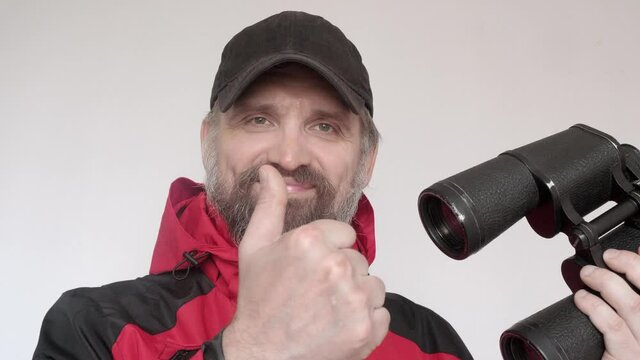 Mature man in red coat demonstrates binoculars, points with a finger an smiles with satisfaction. Man's leisure and hobby.
