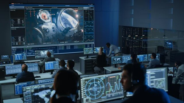 Group of People in Mission Control Center Establish Successful Video Connection on a Big Screen with an Astronaut on Board of a Space Station. Flight Control Scientists Sit in Front of Computers.