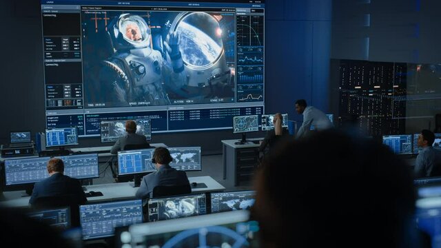 Group of People in Mission Control Center Establish Successful Video Connection on a Big Screen with an Astronaut on Board of a Space Station. Flight Control Scientists Sit in Front of Computers.