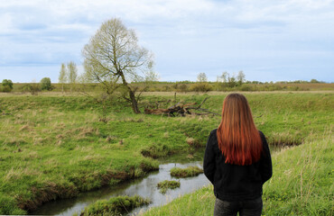 A girl admires the scenery with a lonely tree standing on the bank of a small river covered with bright green grass.