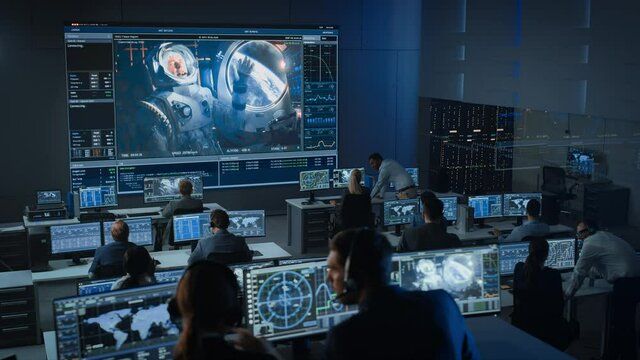 Group of People in Mission Control Center Establish Successful Video Connection on a Big Screen with an Astronaut on Board of a Space Station. Flight Control Scientists Sit in Front of Computers.