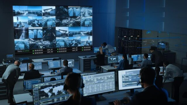 Static Shot of a Group of Professionals Working in a Surveillance Control Center with CCTV Video Footage from an Industrial Factory on a Big Digital Screen. Monitoring Room Employees Sit in Front of C