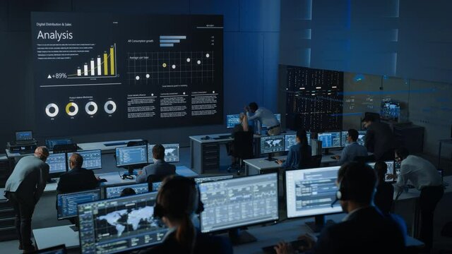 Static Shot of Professional Traders Working in a Modern Monitoring Office with Live Analytics Feed on a Big Digital Screen. Monitoring Room Brokers and Finance Specialists Sit in Front of Computers. 