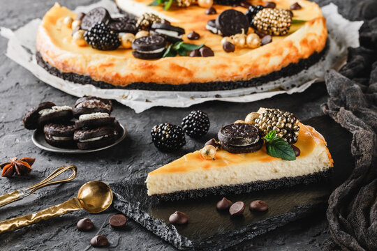 Classic Cheesecake With Caramel, Mini Oreos, Mint Leaves, And Berries On Paper Over Dark Background, Close Up View. Slice Of Cheesecake On Black Rock Plate. Delecious Sweets And Dessert