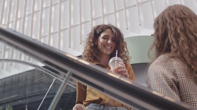 Back view of curly-haired young woman and her female friend with drinks in their hands going up on escalator in shopping center while discussing their purchases